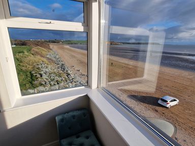 A corner window view overlooking a sandy beach with a white car parked on it and a rocky grassy area at Beach House in Duncannon County Wexford