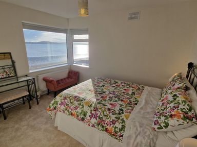 A bedroom with a bed with floral bedding a pink bench a makeup table and windows overlooking the sea at Beach House in Duncannon County Wexford
