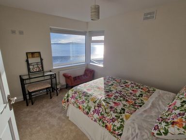 A bedroom with a bed with floral bedding a glass dressing table a pink bench and windows overlooking the sea at Beach House in Duncannon County Wexford
