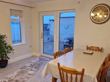 A dining room with a wooden table covered with a patterned tablecloth surrounded by wooden chairs and a large wall clock near a glass door and window at Beach House in Duncannon County Wexford