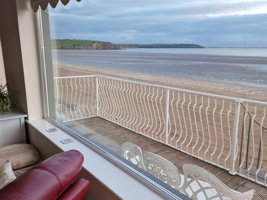 A view through a window showing a sandy beach and sea with a white railing and outdoor chairs on a deck at Beach House in Duncannon County Wexford