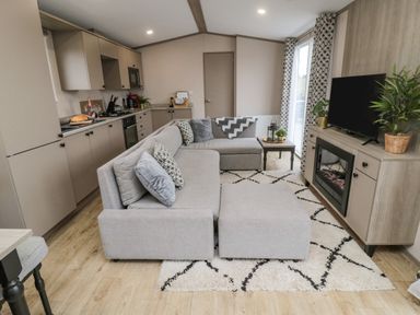An open-plan living room and kitchen with a grey sectional sofa a patterned rug and a TV on a wooden cabinet at Plot 18 Meadow View in Felton