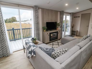 A living room with a gray sofa a television on a cabinet with an electric fireplace and sliding glass doors leading to a balcony at Plot 18 Meadow View in Felton