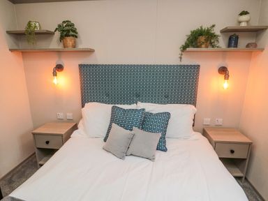 A bedroom with a bed with patterned headboard pillows and bedside tables with shelves and plants on shelves at Plot 18 Meadow View in Felton