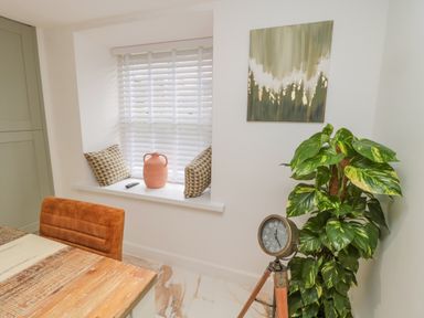 A corner of a room with a wooden table and orange chair a window seat with cushions and a vase a tripod clock and a large leafy plant at Donni Hall Cottage in Beadnell