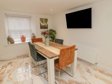 A dining area with a wooden table, four chairs, a window with blinds and cushions, a wall-mounted television, and a vase with flowers at Donni Hall Cottage in Beadnell