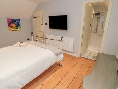 A bedroom with a bed white bedding wooden floor a wall mounted tv an open door to a bathroom with a shower and a colorful wall art at Donni Hall Cottage in Beadnell