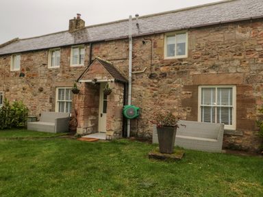 A stone house with two gray benches a front porch with hanging flower pots and a planter on the grass at Donni Hall Cottage in Beadnell