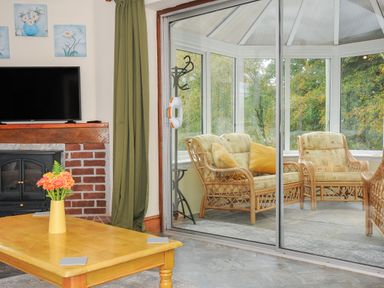 A living room with a coffee table and fireplace adjacent to a conservatory with wicker chairs and large windows at The Wagon House in Lezant near Launceston