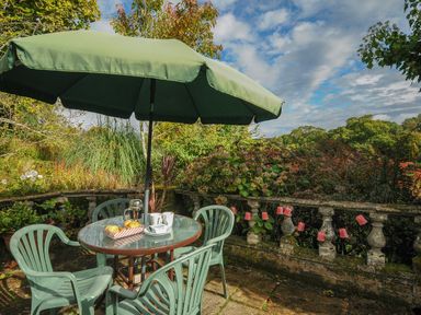 An outdoor patio with a round glass table and four plastic chairs under a green umbrella at The Wagon House in Lezant near Launceston