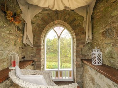 A stone room with a wicker chair a candle in a birdcage and a lantern by a pointed window at The Wagon House in Lezant near Launceston