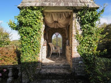 A stone gazebo with a single wicker chair and lanterns surrounded by ivy at The Wagon House in Lezant near Launceston