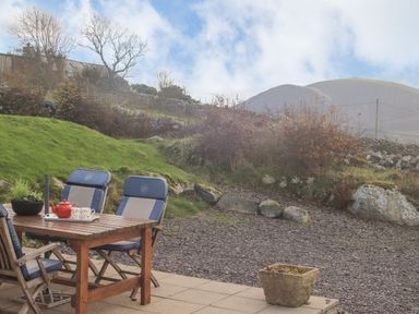 An outdoor patio with a wooden table and cushioned chairs with a teapot and cups on the table and a hillside with rocks and bushes in the background at Beech Tree Cottage in Waunfawr