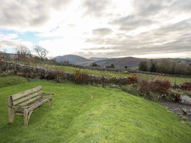A wooden bench on a grassy area with stone walls and hills in the background at Beech Tree Cottage in Waunfawr