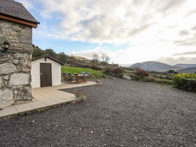 An outdoor patio area with a small shed and a wooden table with chairs on a tiled surface next to a gravel ground and hills in the background at Beech Tree Cottage in Waunfawr
