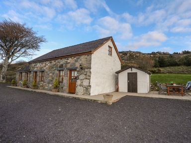 A stone cottage with wooden doors and windows next to a small white shed and outdoor seating area at Beech Tree Cottage in Waunfawr