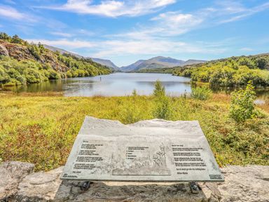 A landscape view of a lake with surrounding greenery and hills with an information plaque in the foreground at Beech Tree Cottage in Waunfawr