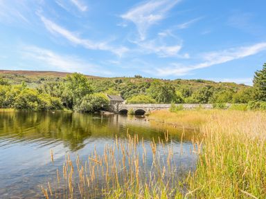 A stone bridge over a river surrounded by trees and grass with hills and a house in the background at Beech Tree Cottage in Waunfawr