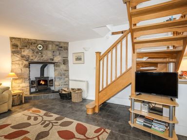 A living room with a stone fireplace wood stove wooden stairs and a television on a stand at Beech Tree Cottage in Waunfawr