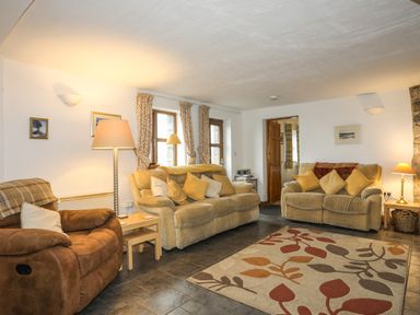 A living room with two beige sofas, a brown armchair, a floor lamp, side tables, patterned curtains, and a rug with leaf designs at Beech Tree Cottage in Waunfawr