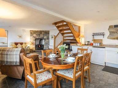 A dining area with a wooden table set with dishes and glasses next to a kitchen and a living room with a stone fireplace and wooden staircase at Beech Tree Cottage in Waunfawr