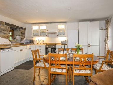 A kitchen and dining area with wooden chairs around a table set for a meal and white cabinets in the background at Beech Tree Cottage in Waunfawr