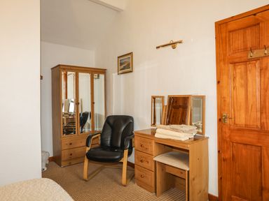 A room with a wooden wardrobe, a black chair, a wooden dressing table with a mirror and folded towels on top at Beech Tree Cottage in Waunfawr