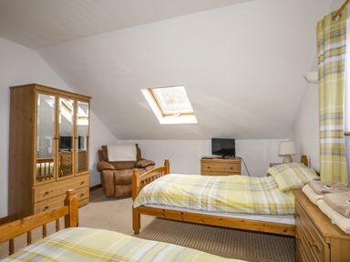 A bedroom with two single beds a wooden wardrobe a brown armchair a wooden chest of drawers with a small television and a lamp and a skylight window at Beech Tree Cottage in Waunfawr