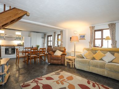 An open plan kitchen and living room with a dining table wooden chairs a beige sofa an armchair two table lamps and a patterned rug at Beech Tree Cottage in Waunfawr