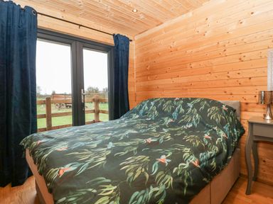 A bedroom with a bed covered with a leaf and bird patterned duvet next to a nightstand and glass door with dark curtains at The Lodge at Crossroads Farm in Middlezoy