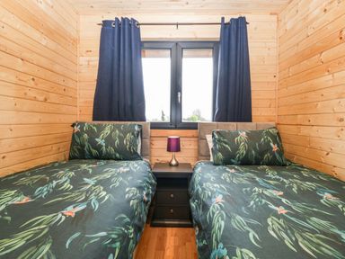 A wooden bedroom with two single beds green leaf patterned bedding a black nightstand and blue curtains at The Lodge at Crossroads Farm in Middlezoy