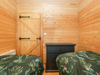 A bedroom with wooden walls a wooden door a black dresser and two beds with green patterned bedding at The Lodge at Crossroads Farm in Middlezoy