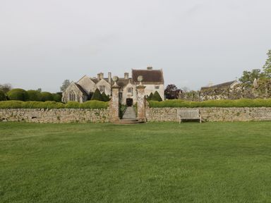 A large house with stone walls and gate surrounded by manicured bushes and a bench on the lawn at The Lodge at Crossroads Farm in Middlezoy