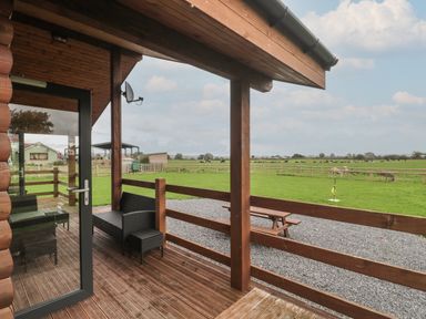 A wooden porch with black outdoor furniture and a picnic table overlooking a fenced green field with grazing animals at The Lodge at Crossroads Farm in Middlezoy