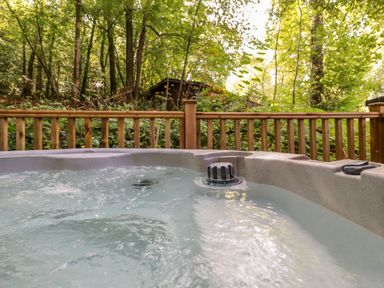 An outdoor hot tub with water bubbling surrounded by a wooden railing and trees at Woodland Retreat Skiptory Howe 41A White Cross Bay near Windermere