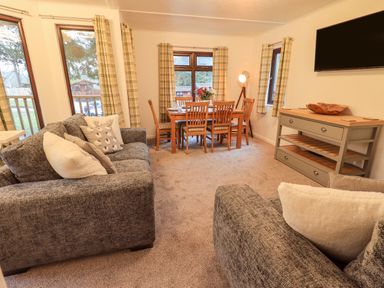 A living room with grey sofas, wooden dining table and chairs, a sideboard, and a wall mounted TV at Woodland Retreat Skiptory Howe 41A White Cross Bay near Windermere