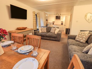 A living room with a dining table set with plates and glasses two sofas with cushions a wall-mounted TV and a view of the kitchen at Woodland Retreat Skiptory Howe 41A White Cross Bay near Windermere