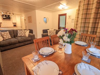 A dining table set with plates glasses and a vase of flowers next to a living area with sofas and cushions at Woodland Retreat Skiptory Howe 41A White Cross Bay near Windermere
