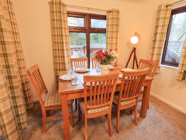 A dining room with a wooden table and six chairs set with plates and glasses with windows and plaid curtains at Woodland Retreat Skiptory Howe 41A White Cross Bay near Windermere