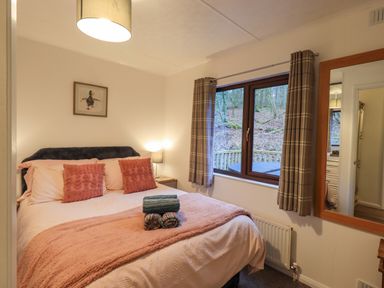 A bedroom with a bed with pink bedding and pillows a bedside table with a lamp a window with plaid curtains and a wooden framed mirror on the wall at Woodland Retreat Skiptory Howe 41A White Cross Bay near Windermere
