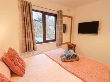 A bedroom with a bed pillows folded towels on the bed a window with curtains a wall mounted tv and a small wooden table at Woodland Retreat in Skiptory Howe 41A White Cross Bay near Windermere