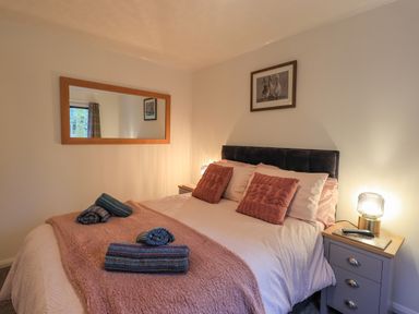 A bedroom with a bed with pink blankets and pillows bedside tables with lamps a mirror and a framed picture on the wall at Woodland Retreat Skiptory Howe 41A White Cross Bay near Windermere