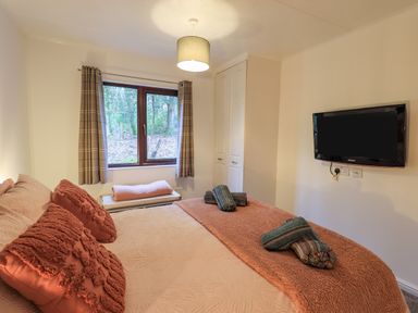 A bedroom with a double bed orange pillows and blanket a window with curtains a wall mounted television and a wardrobe at Woodland Retreat Skiptory Howe 41A White Cross Bay near Windermere