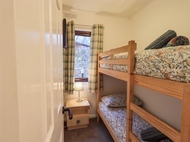A small bedroom with wooden bunk beds and a nightstand with a lamp next to a window with plaid curtains at Woodland Retreat in Skiptory Howe 41A White Cross Bay near Windermere