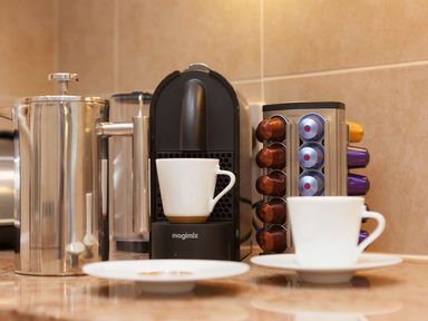 A coffee machine with a white cup a metal French press and a capsule holder with coffee pods on a kitchen counter at Surf and Sand in Carbis Bay