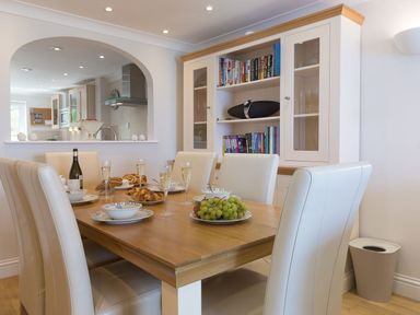 A dining room with a wooden table set with plates glasses and food six white chairs and a cupboard with books and glassware