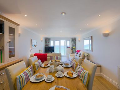 A dining table set with breakfast items and chairs with cushions in a living room with sofas a television and sea view at Surf and Sand in Carbis Bay
