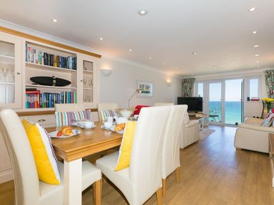 A dining area with a wooden table and chairs and a living room with a television and sofas overlooking the sea at Surf and Sand in Carbis Bay