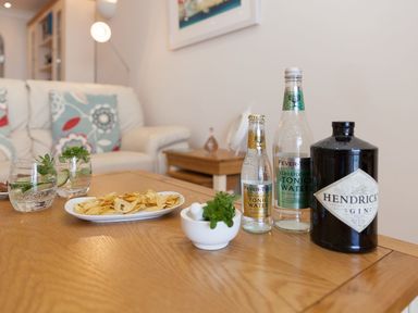 A wooden table with gin bottle tonic water glasses and a plate of chips in a living room at Surf and Sand in Carbis Bay