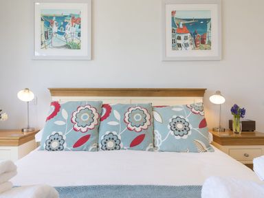A bedroom with a white bed with patterned pillows two bedside tables with lamps and flowers and two framed paintings on the wall at Surf and Sand in Carbis Bay